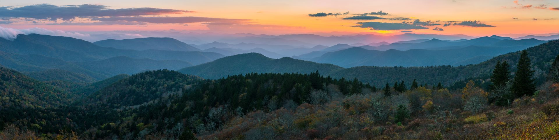 A panoramic view of rolling, tree-covered mountains at sunset, with soft pink and orange clouds above the horizon.