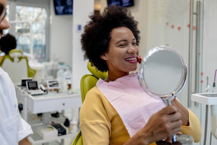 A woman is sitting in a dental chair looking at her teeth in a mirror.