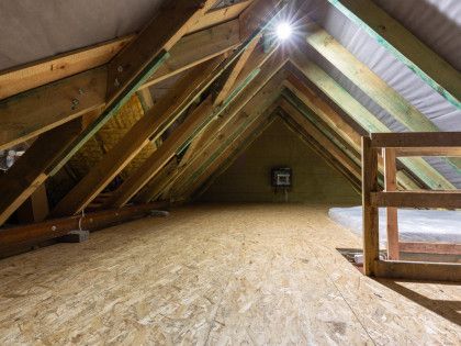 Attic interior with exposed wooden beams, plywood floor, and a bright light at the peak