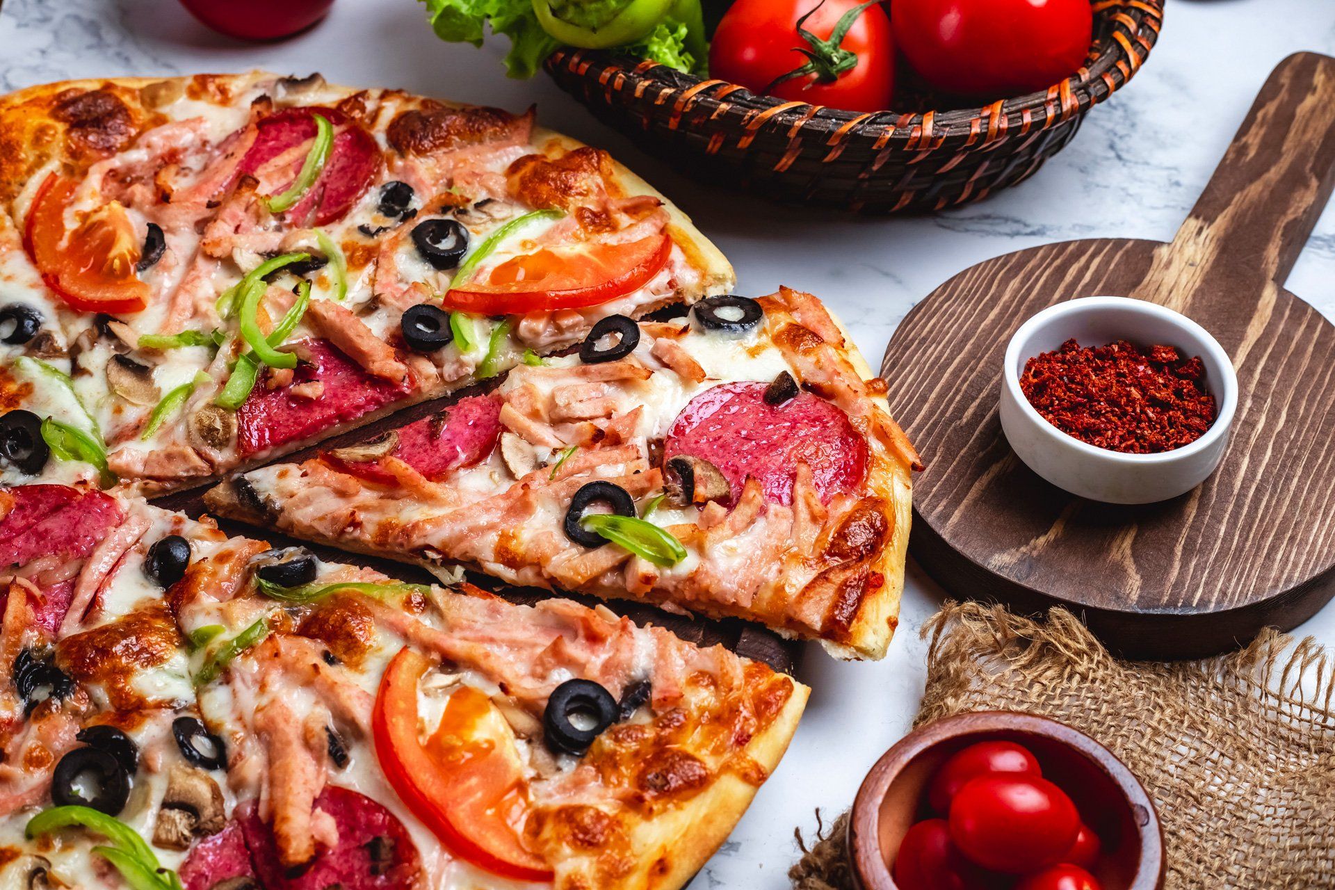 A pizza with pepperoni , olives , tomatoes and other toppings on a table.