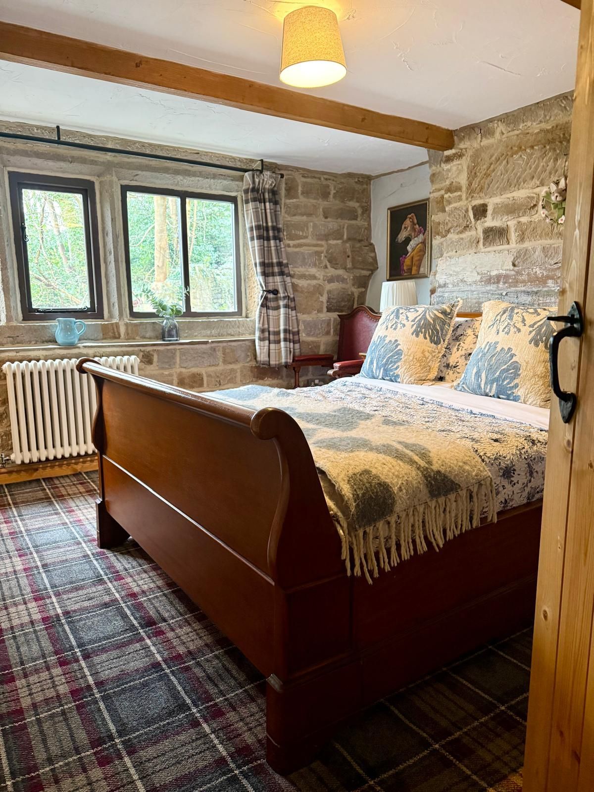 Cozy stone bedroom with wooden bed, plaid rug, and windows overlooking greenery