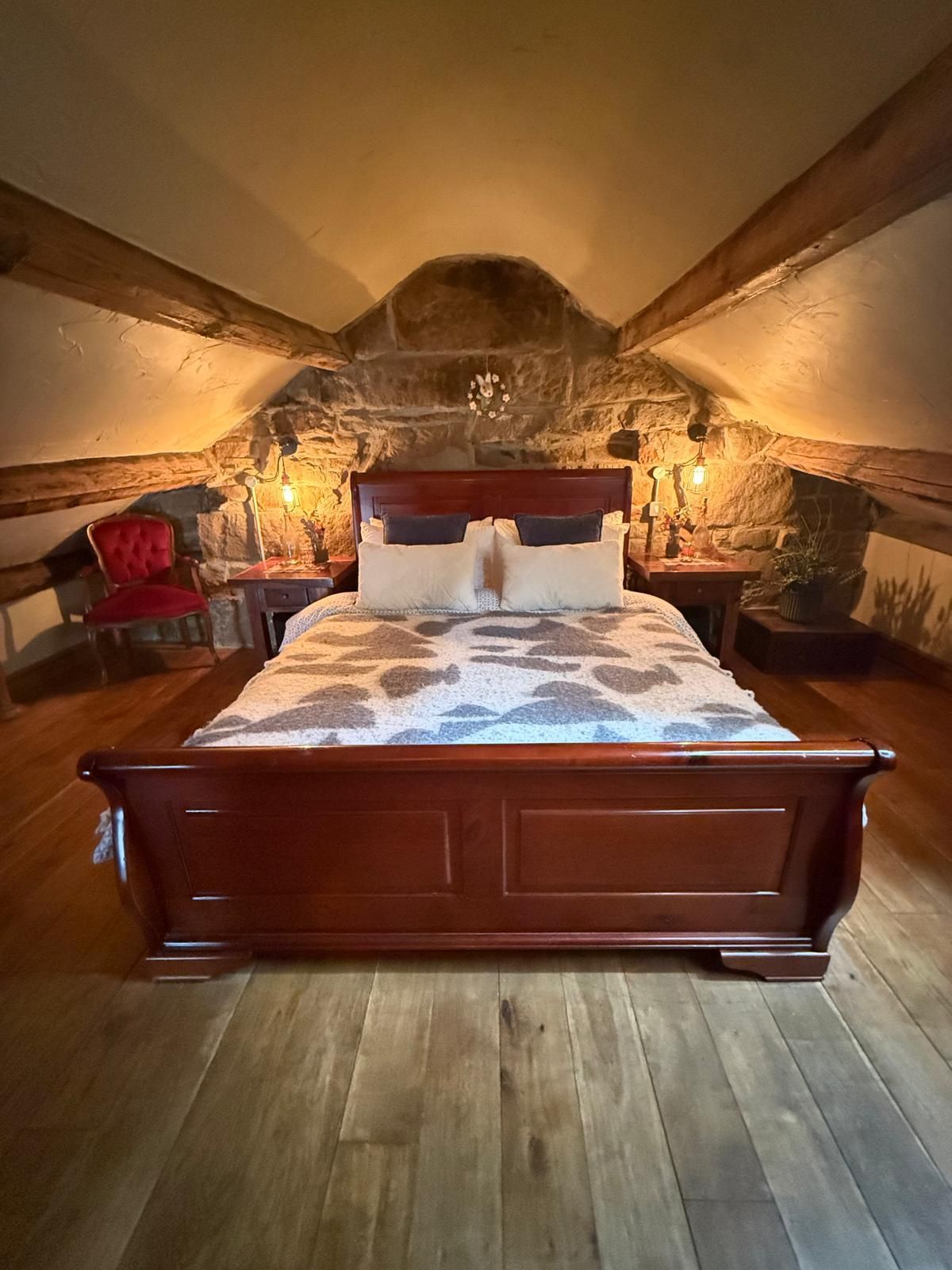 Cozy attic bedroom with wood beams, a wooden bed, warm lamp light, and rustic stone wall backdrop