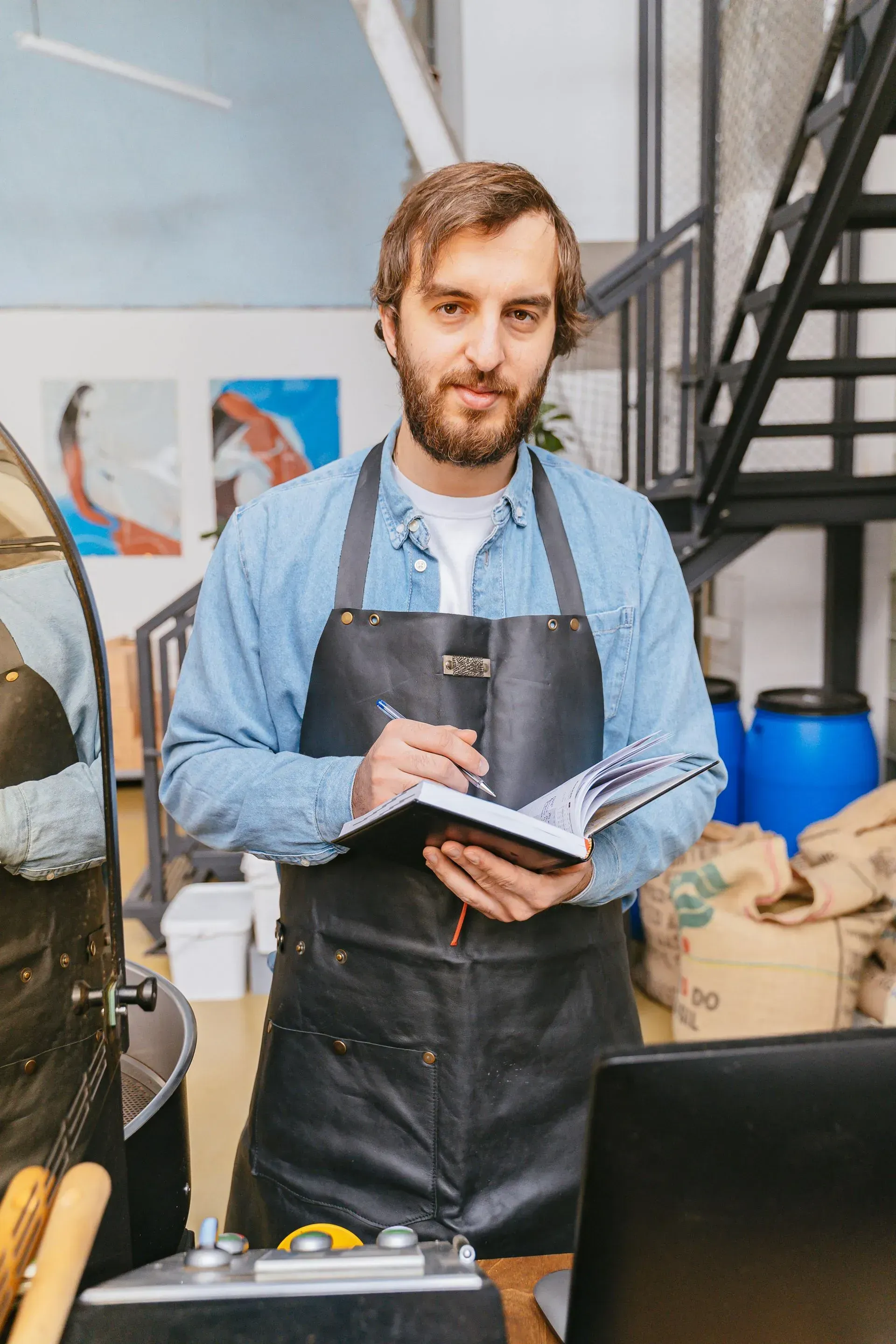 Man in a blue shirt and apron holding a notebook in a workshop beside a laptop and stairs