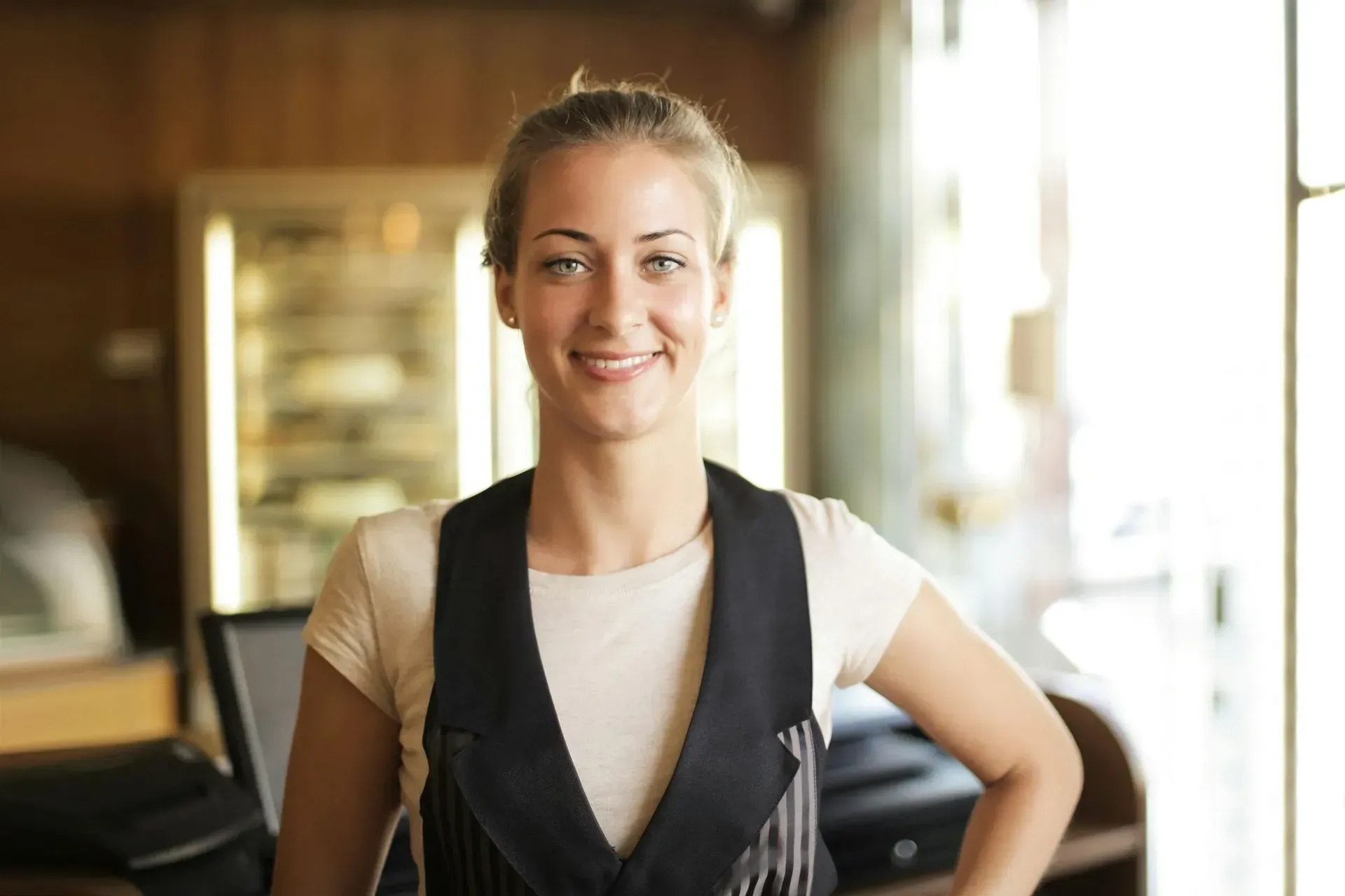 Smiling woman standing indoors near a bright window, wearing a white shirt and dark vest.