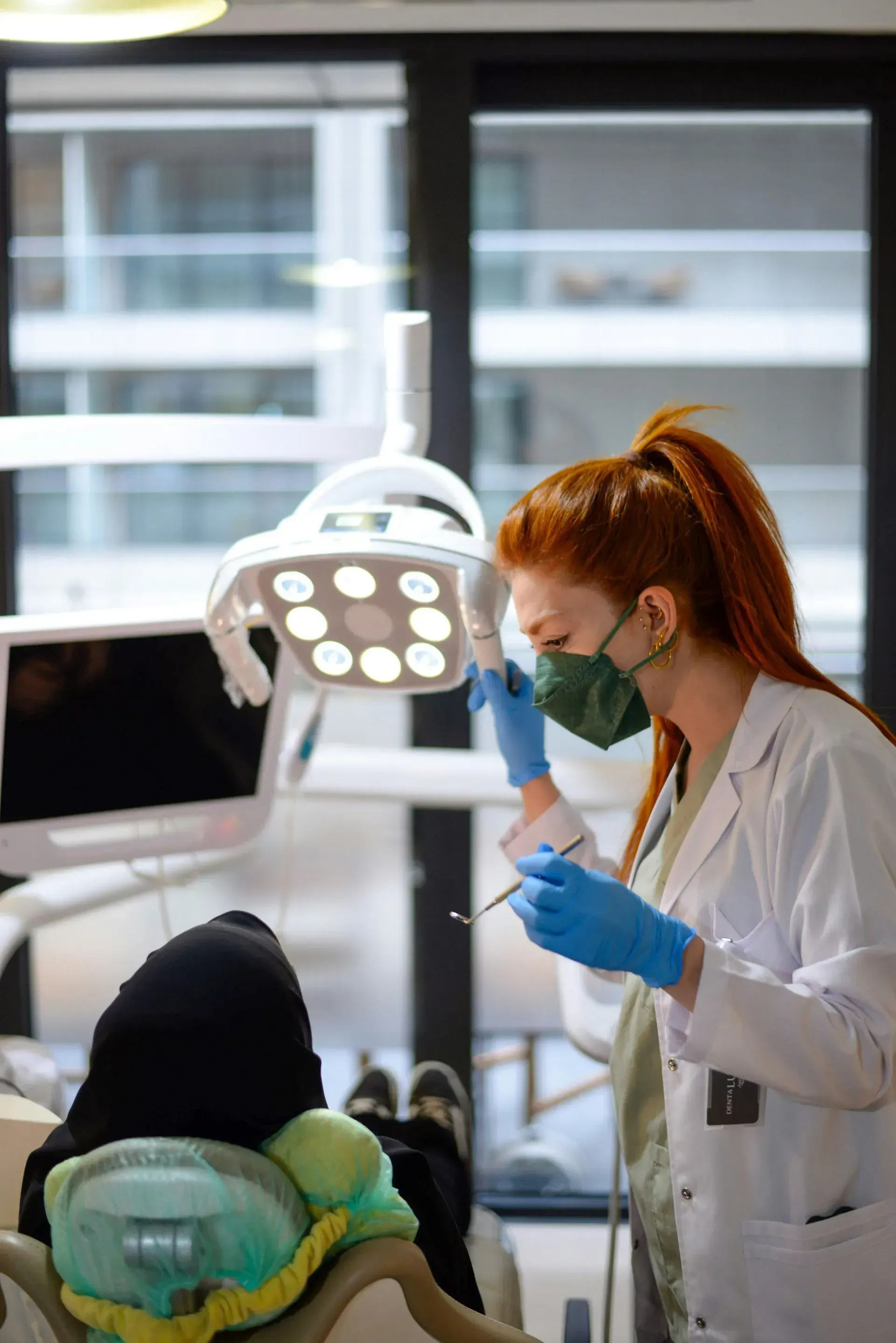 Dentist in white coat and blue gloves examines a patient in a dental chair under bright exam light