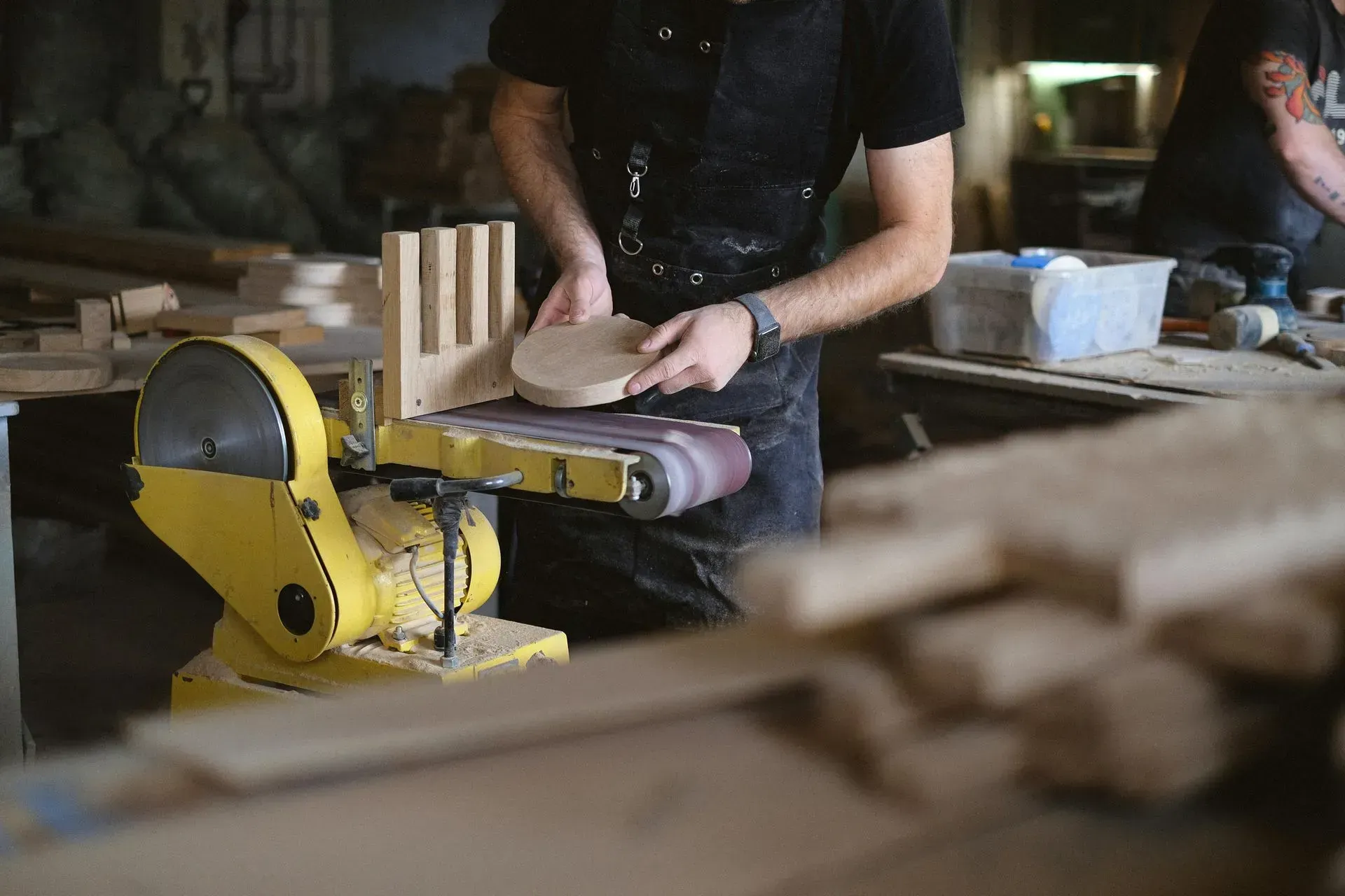Person sanding wood at a workbench beside a yellow power sander in a workshop