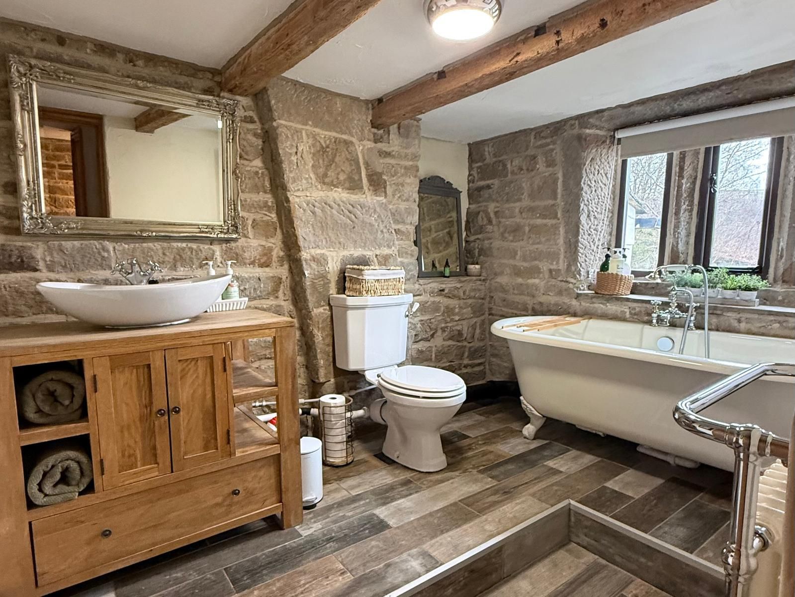 Rustic stone bathroom with wood beams, vanity, tub, toilet, and window light