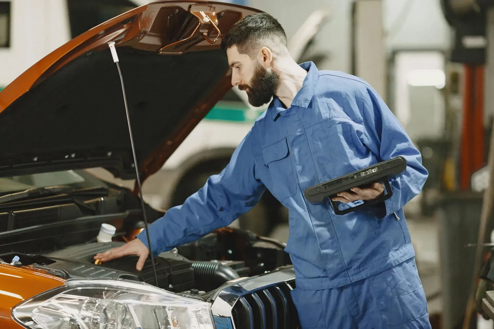 Mechanic in blue coveralls inspecting a car engine with the hood open in a garage