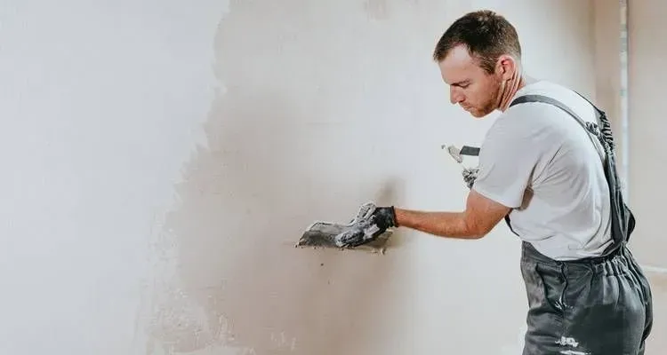 Person plastering a wall with a trowel, wearing white shirt and gray overalls in a bright room