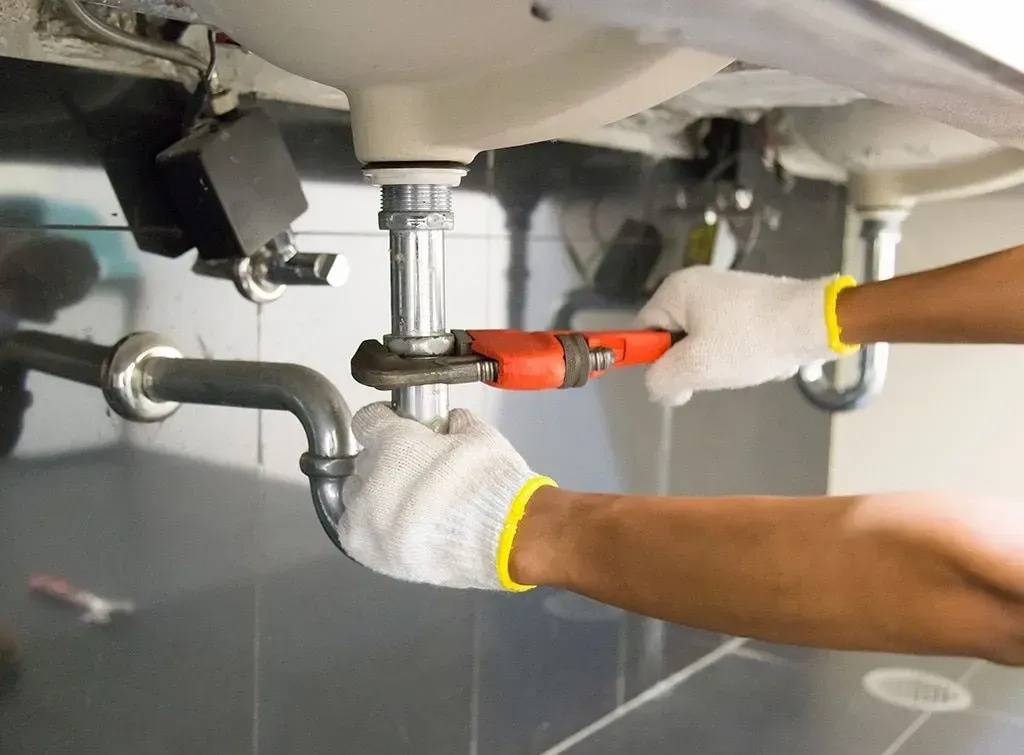 Gloved hands use a red pipe wrench to tighten plumbing under a sink