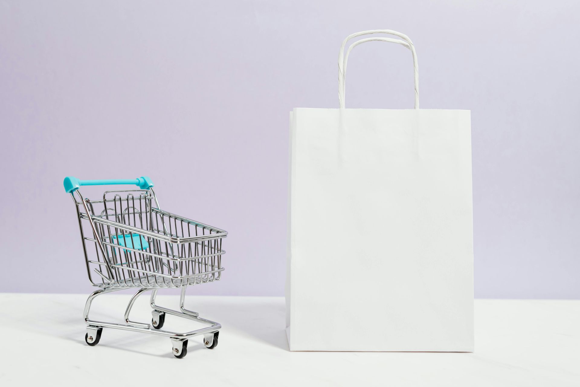 Mini shopping cart and white paper bag on a table against a light purple background.