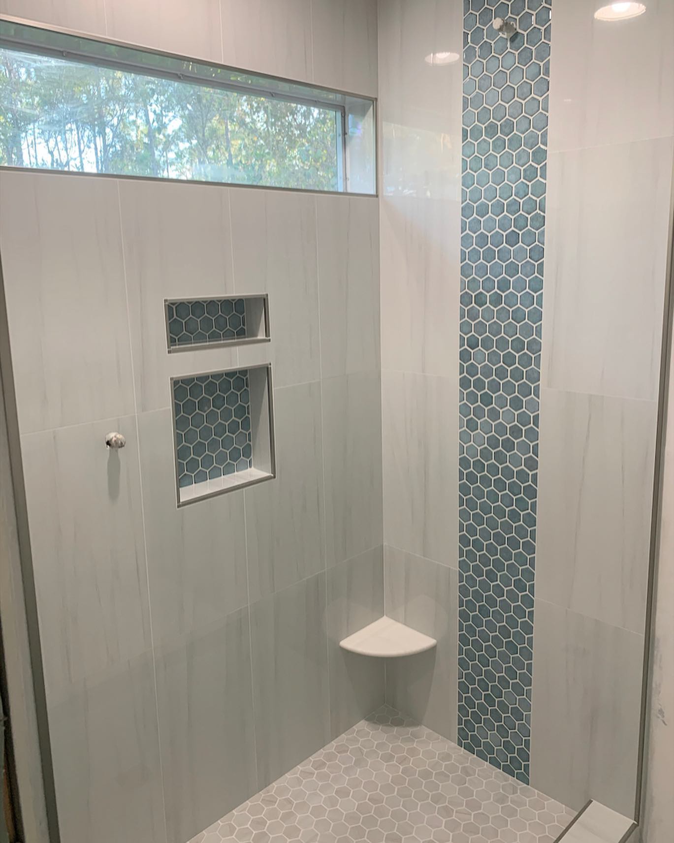 Modern tiled bathroom with glass shower, mosaic accent strip, small window, and corner shelf.