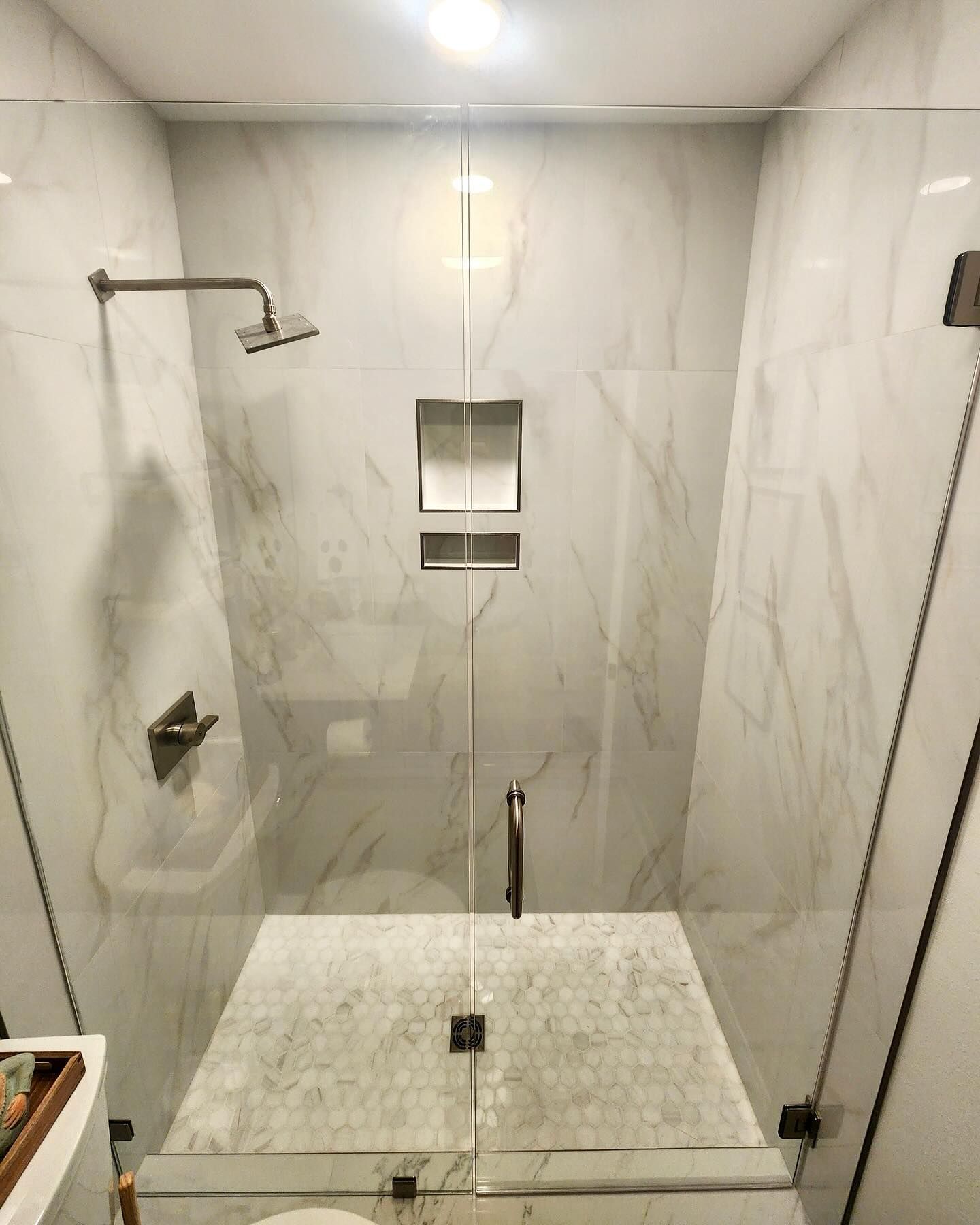 Modern marble-tiled shower stall with glass door, rain showerhead, built-in niche, and pebble-textured floor.