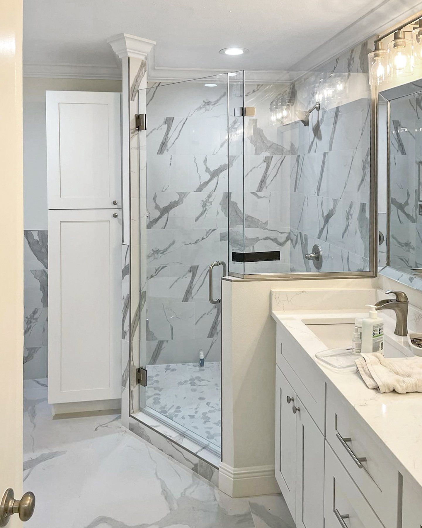 Bright bathroom with glass shower, marble-pattern tile, white vanity, and tiled floor.