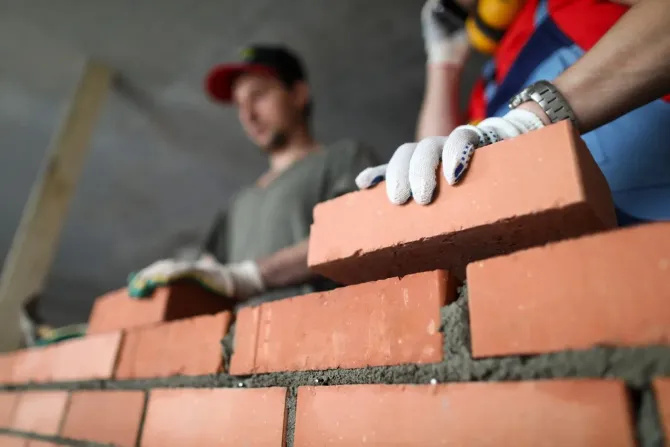 Gloved workers laying orange bricks on a wall at a construction site