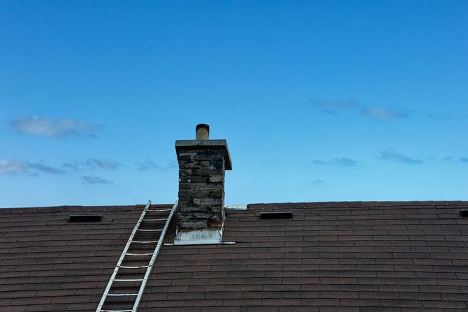 Rooftop with a stone chimney and a ladder leaning against it under a blue sky.