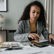 Woman at a desk counting money with a calculator beside a laptop in a home office