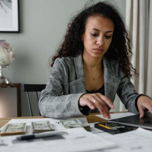 Woman at a desk counting money with a calculator beside a laptop in a home office