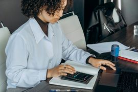 Person typing on a calculator at a desk with a laptop and notebook nearby