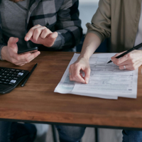 Two people at a table reviewing documents, one using a smartphone beside a calculator and coffee mug