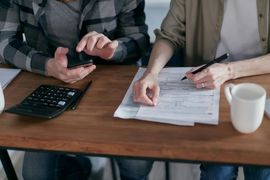 Two people reviewing paperwork at a wooden table with a calculator, pen, and coffee mug