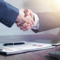 Two people in suits shaking hands over a desk with a tablet and papers