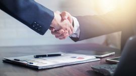 Business handshake over a desk with documents, pen, and laptop in a bright office setting