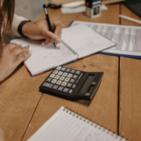 Person writing in a notebook beside a calculator, papers, and clipboard on a wooden desk