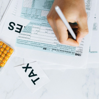 Hand writing on a tax form beside a calculator and orange pills on a white desk