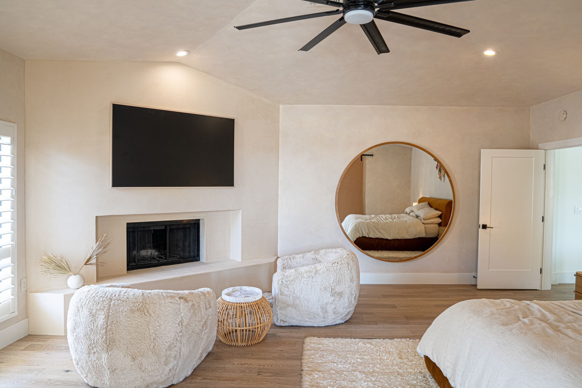 Bright bedroom with a fireplace, wall TV, round mirror, and neutral decor in beige tones