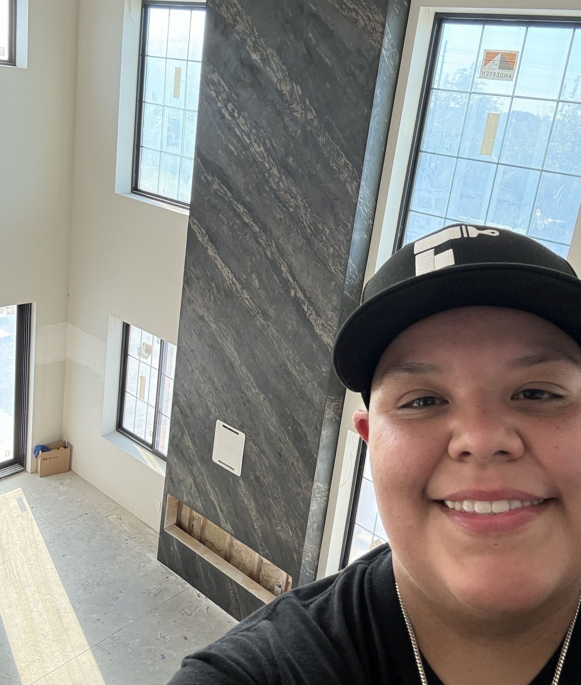 Smiling person in black cap taking a selfie in a bright stairwell with large windows and a dark accent wall