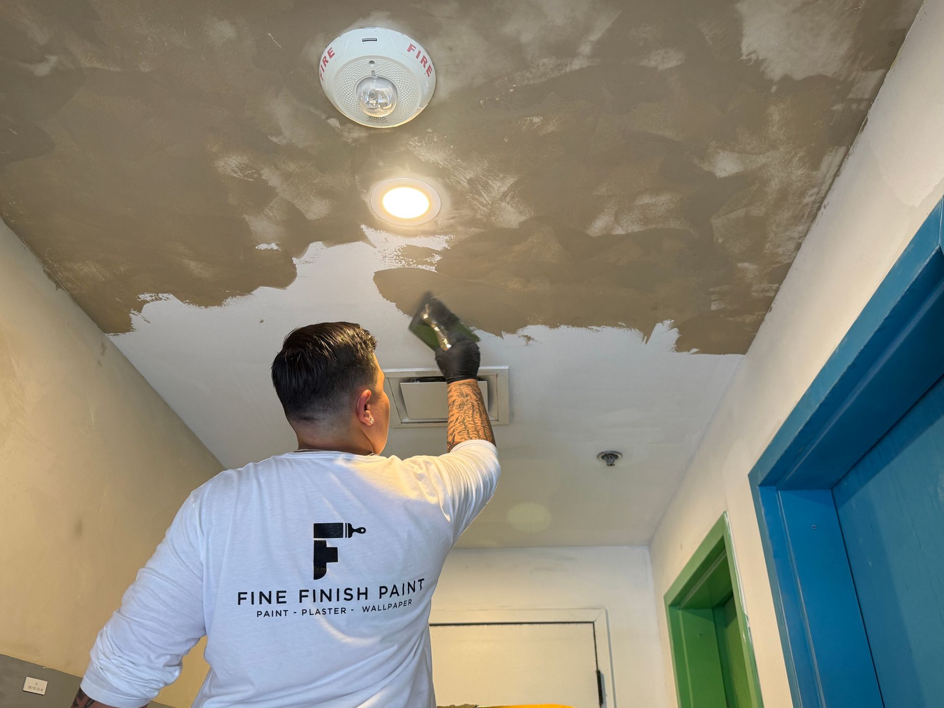 Worker using a sprayer on a patched ceiling in a hallway with blue and green doors