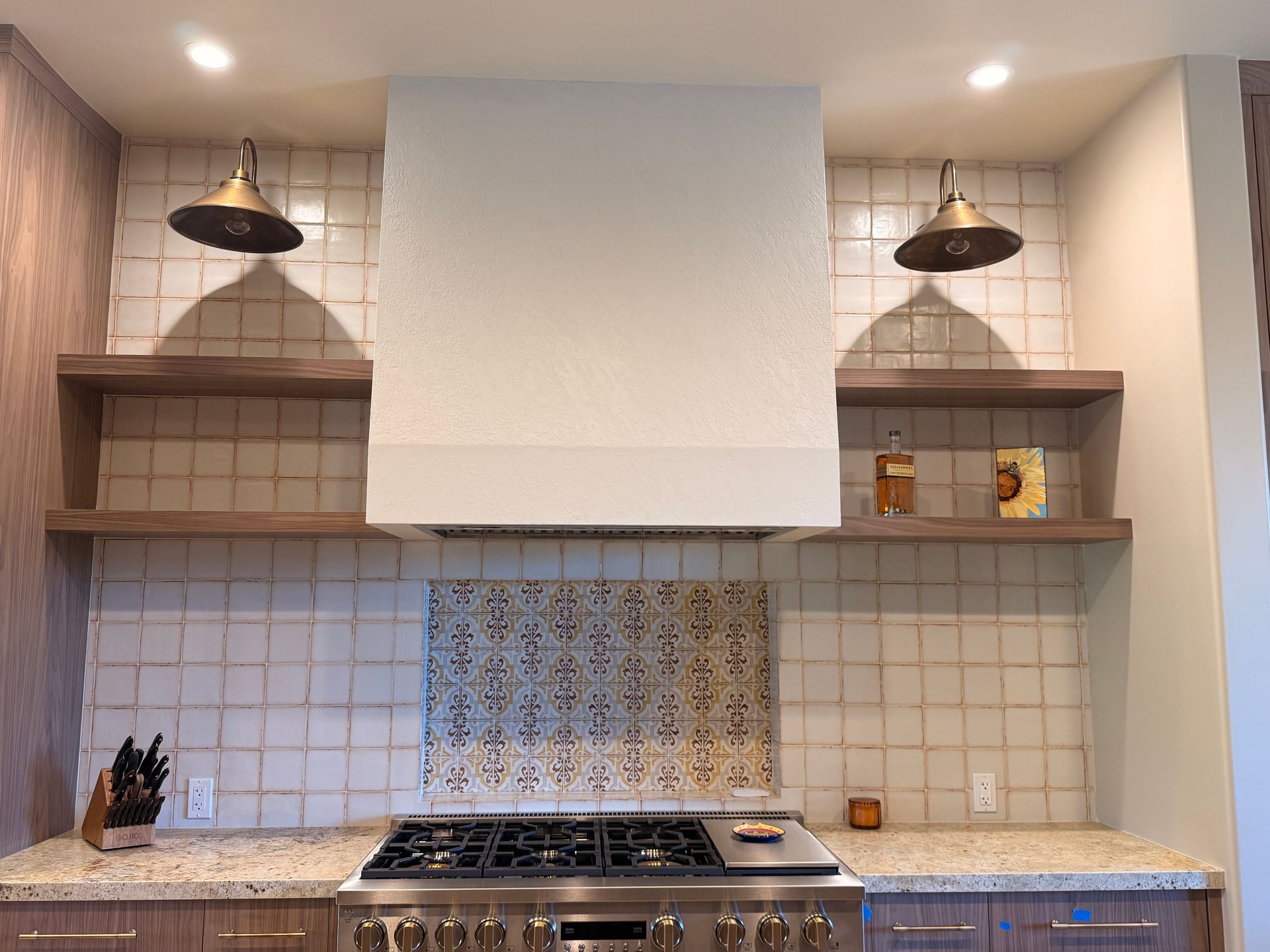 Modern kitchen with white tile backsplash, stainless stove, wood shelves, and hanging black pendant lights