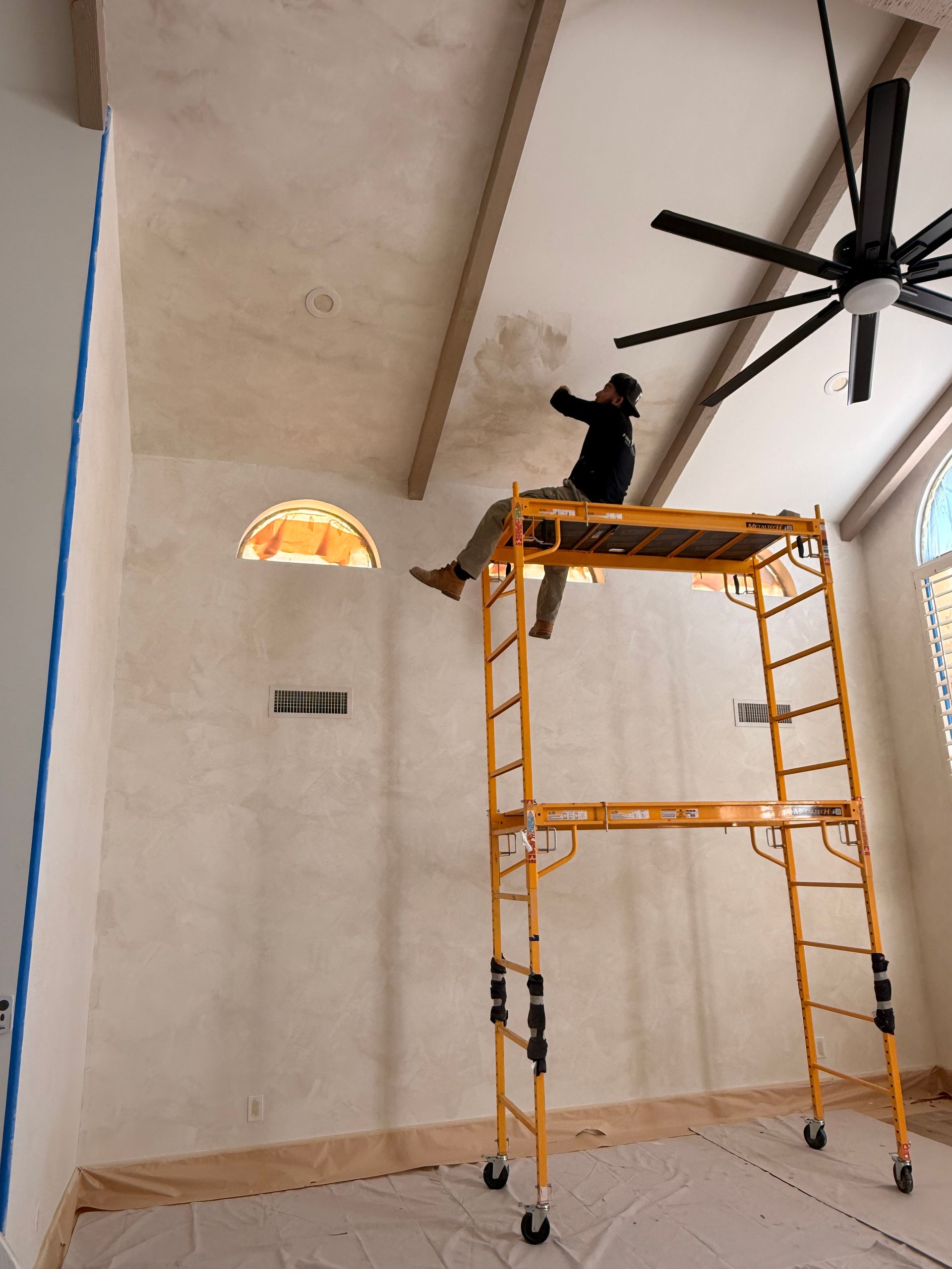 Person on a yellow scaffold installing a large black ceiling fan in a tall, unfinished room.