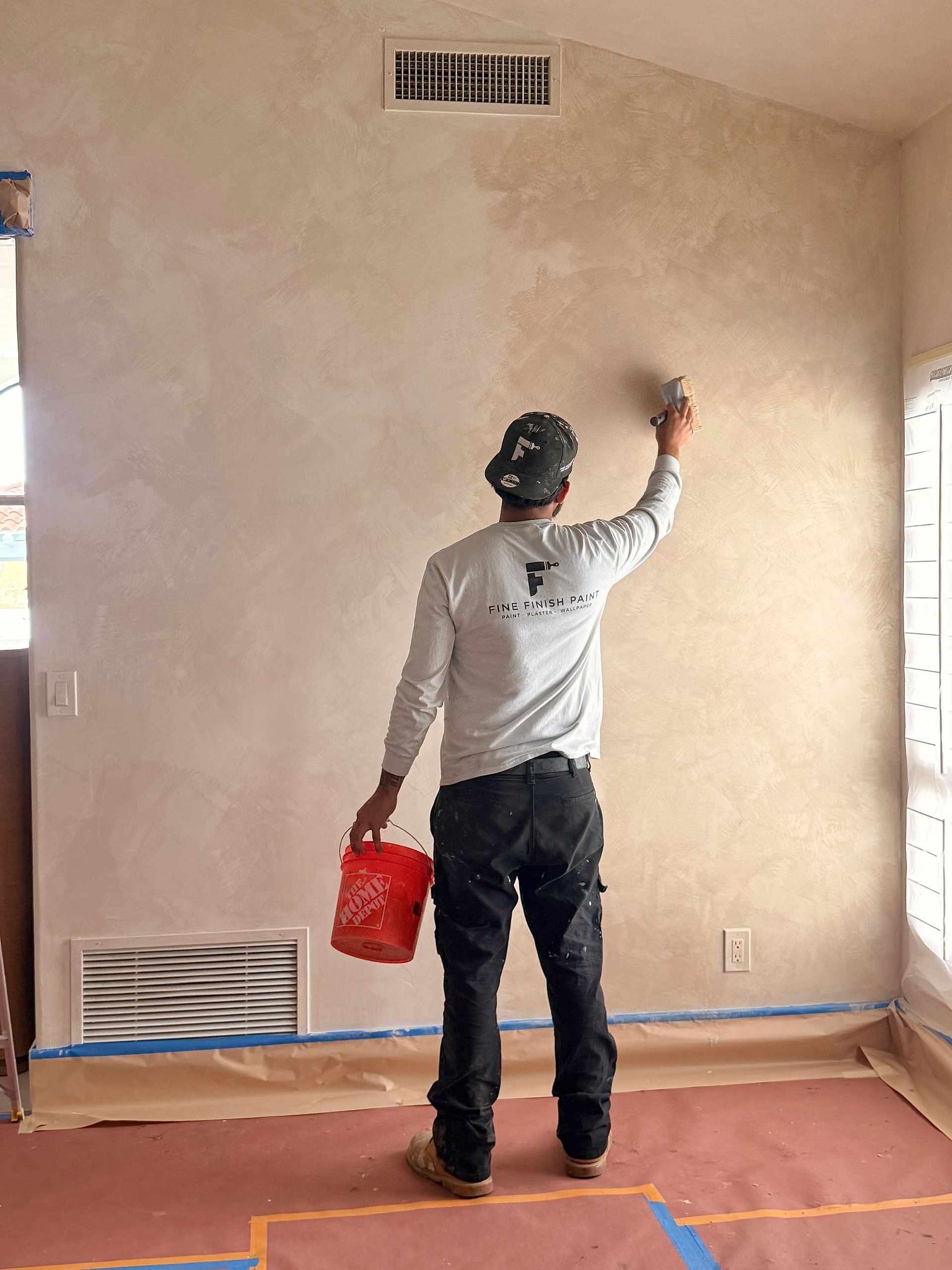 Person painting a beige wall with a roller while holding a red bucket in an unfinished room