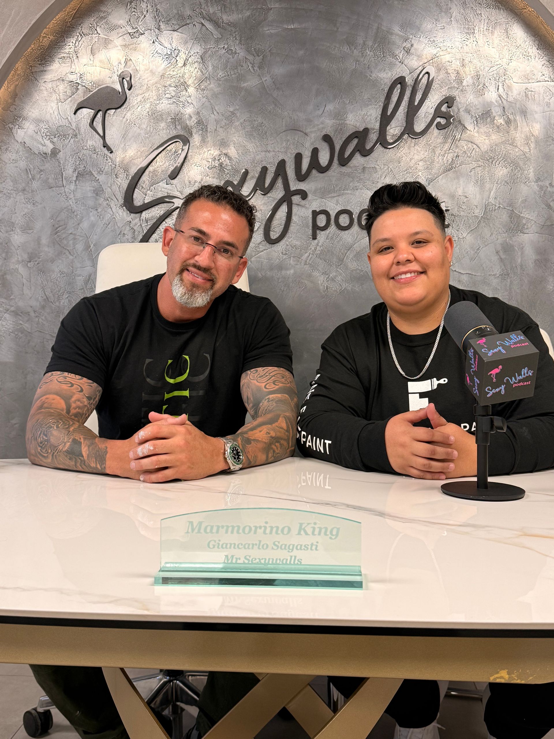 Two people seated behind a table at a wall sign, smiling and holding microphones.