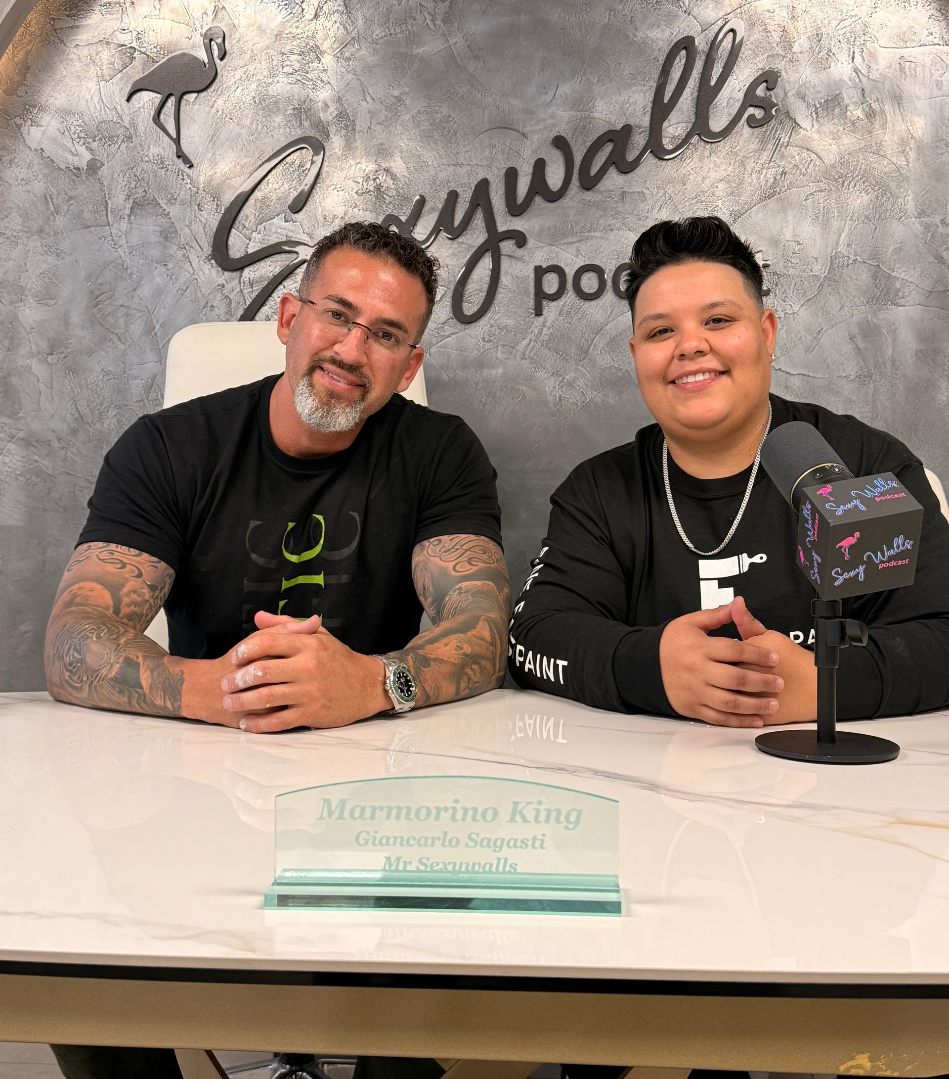 Two hosts smiling at a table with a microphone and nameplate in front of a branded wall.