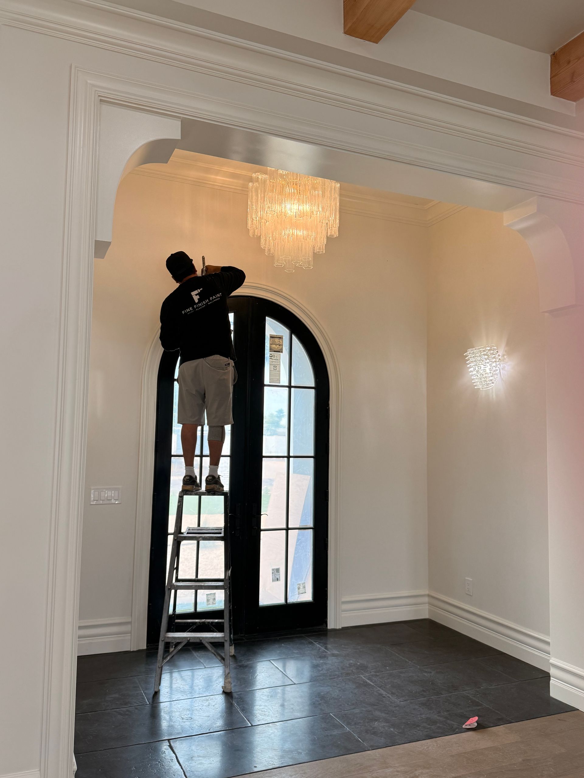 Person on a ladder installing a chandelier in a white room with arched doorway and black floor.