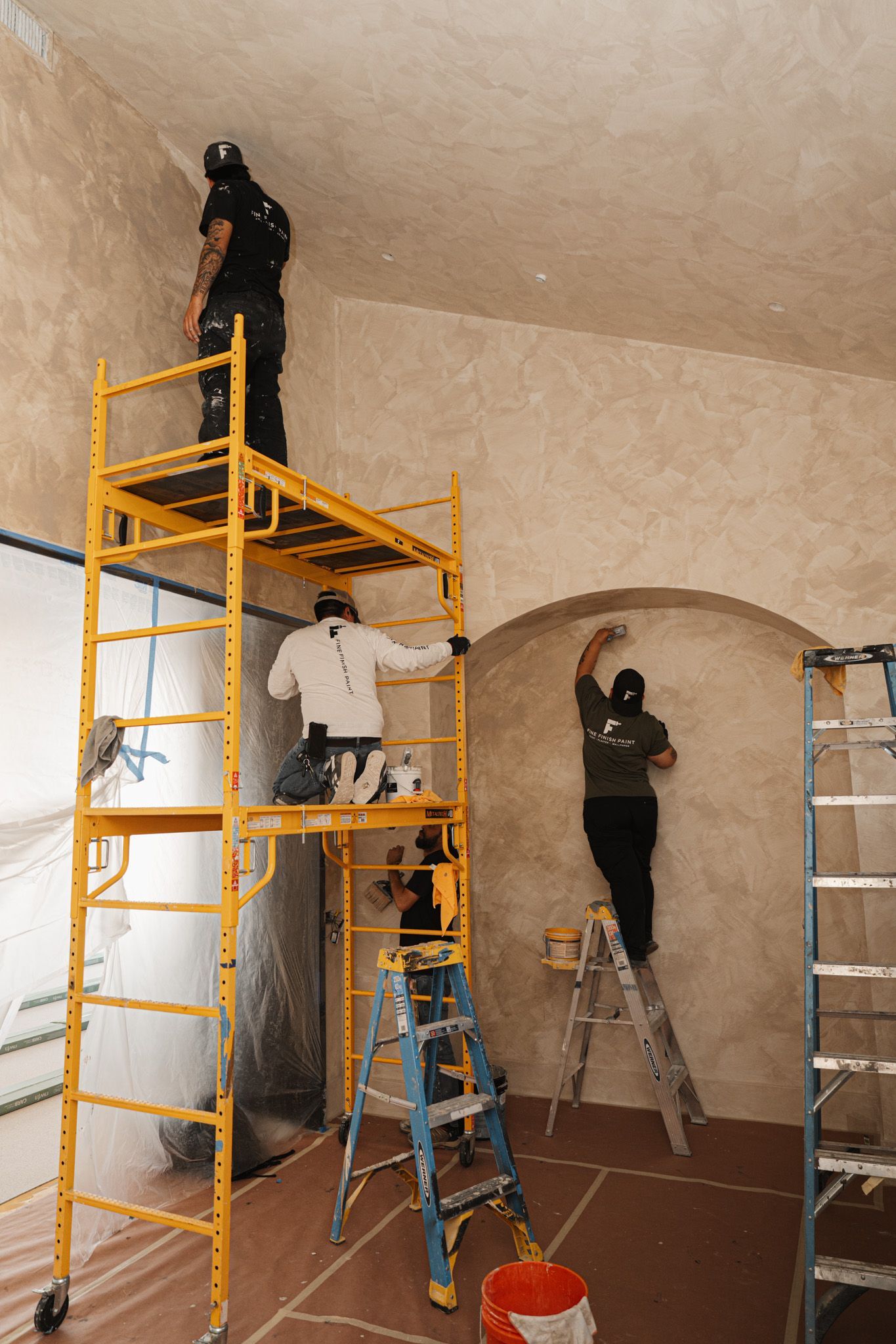 Workers on ladders and scaffolding painting a room with high ceilings and arched walls.