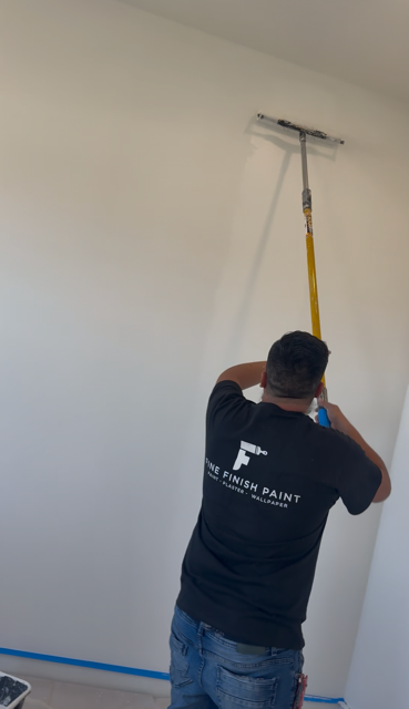 Man in black shirt painting a white wall with a long-handled roller indoors