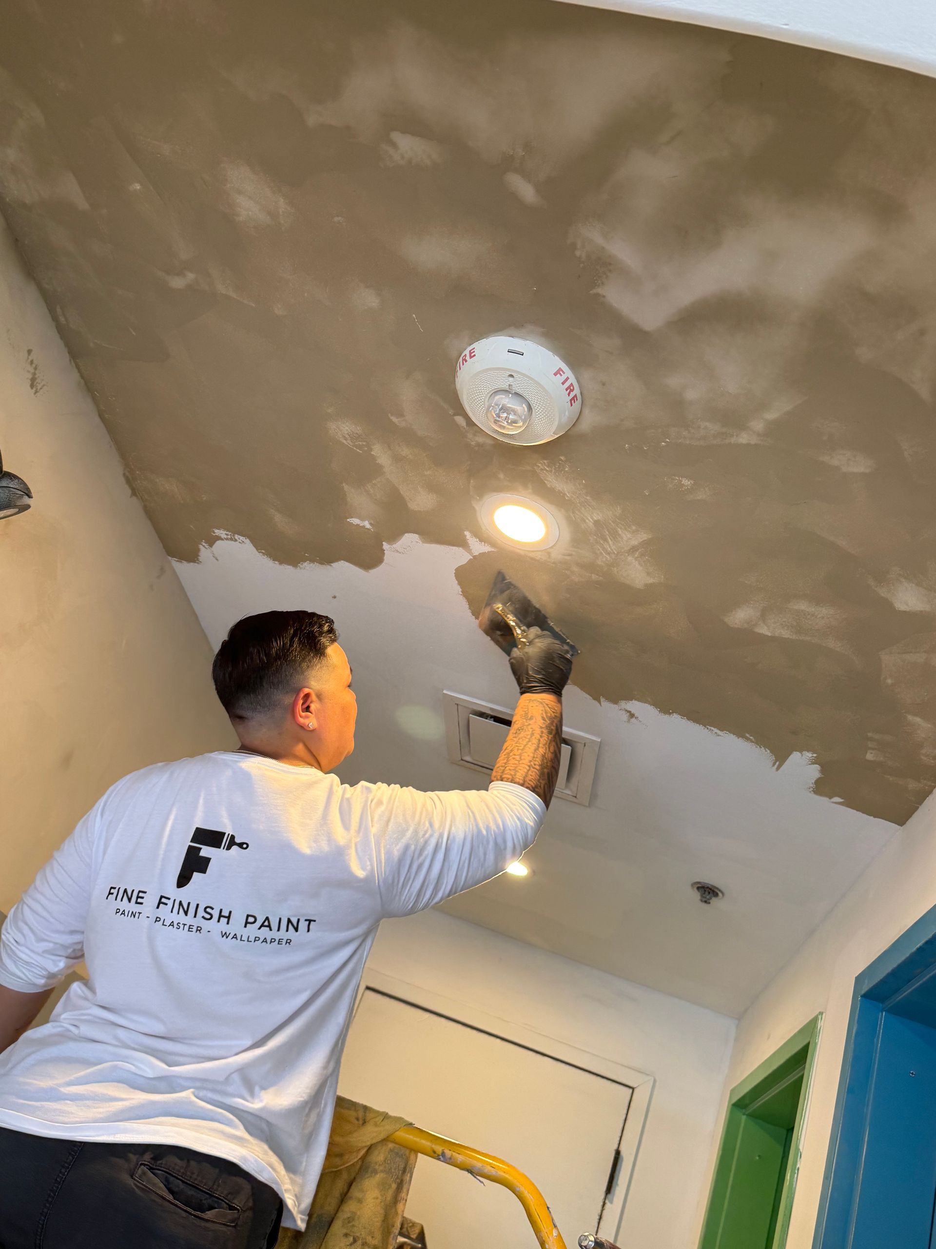 Worker plastering a ceiling in a hallway with a trowel, near a light fixture and smoke detector.
