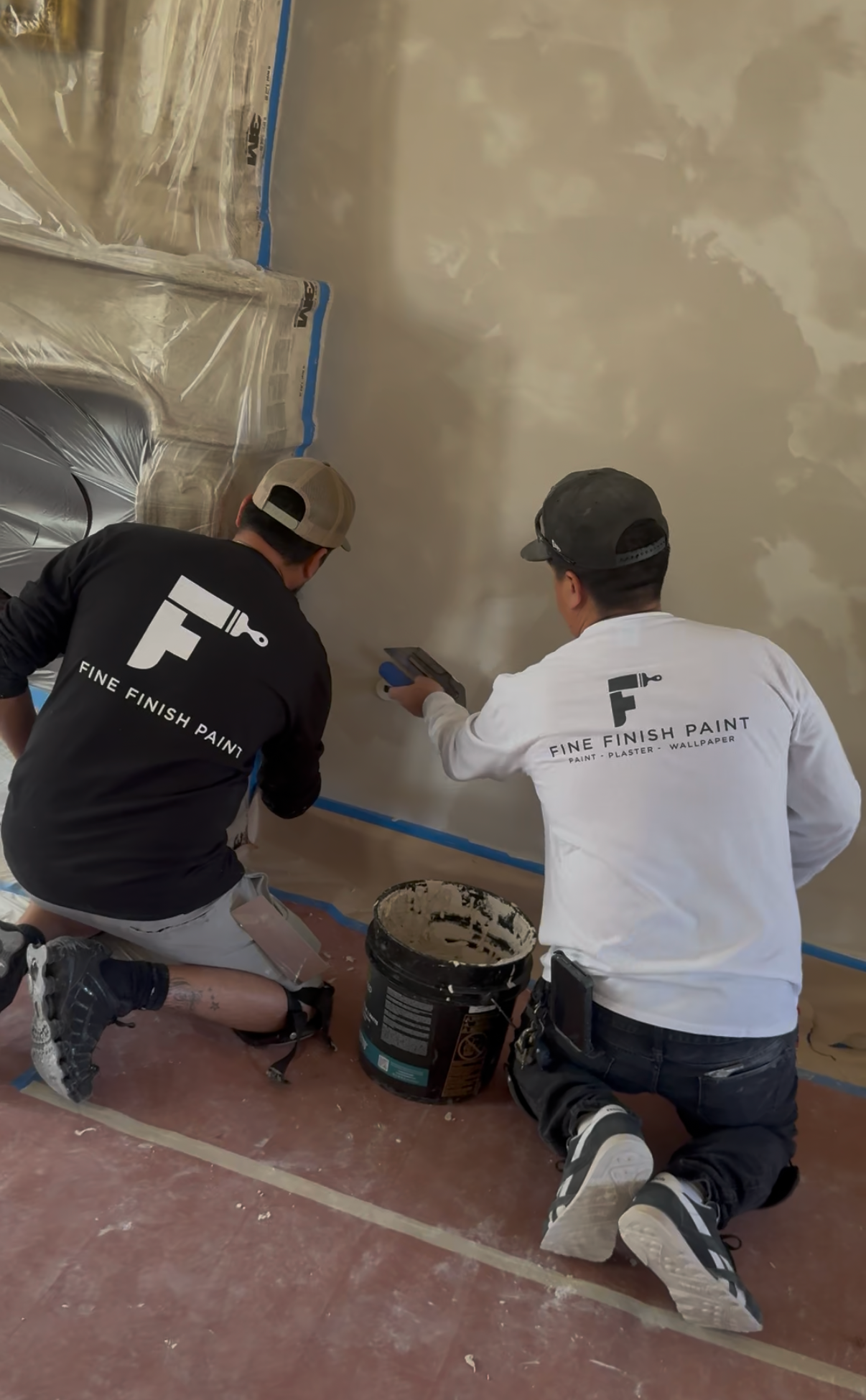 Two painters kneel and brush blue tape along a wall in a room being prepped for painting.