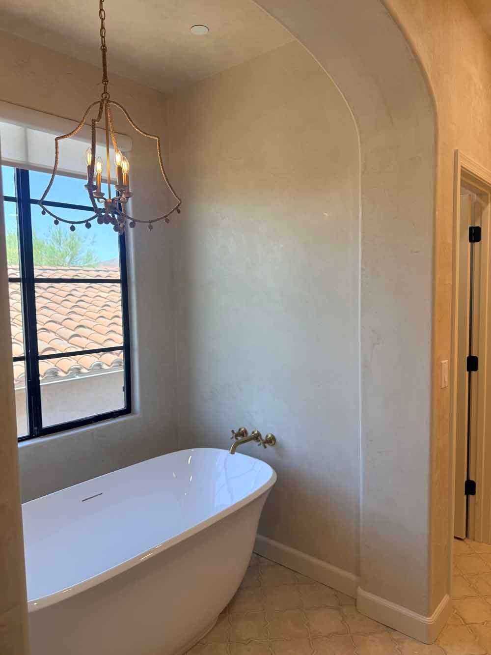 Modern bathroom with freestanding tub, arched alcove, window, and hanging chandelier