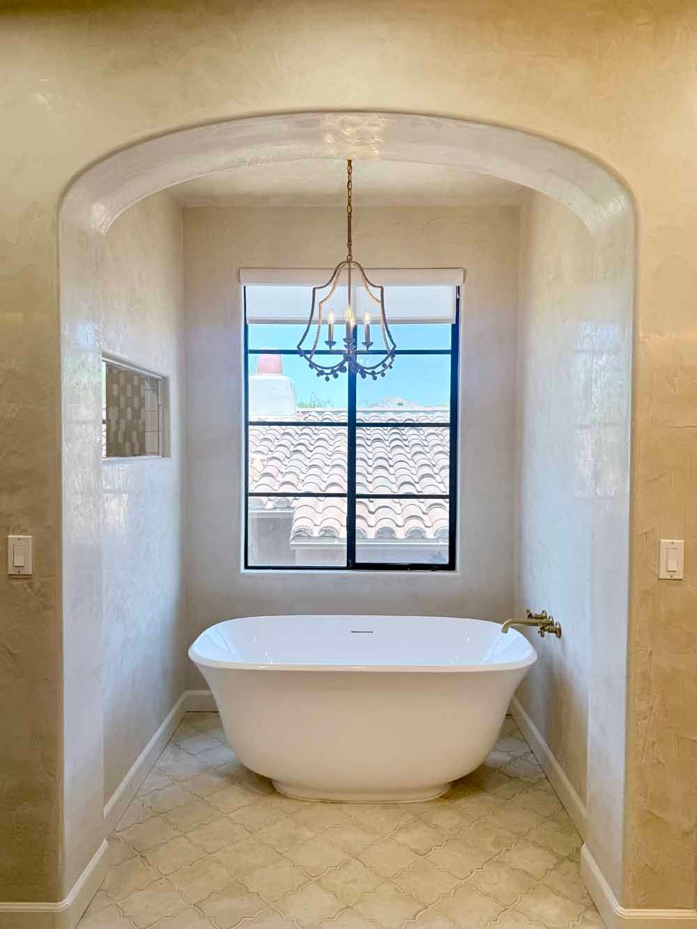 A bright bathroom with a freestanding white tub beneath a window and chandelier.