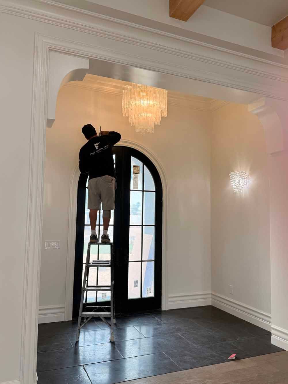Worker on a ladder painting a white arched doorway in a bright room with dark tile flooring.