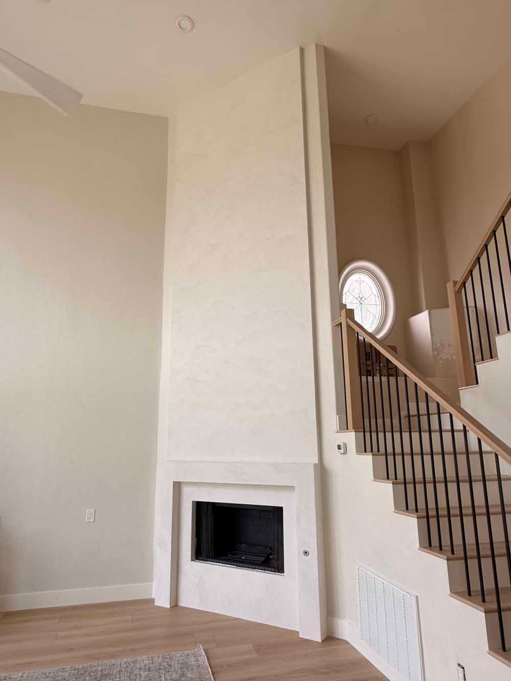Living room with tall white fireplace beside a staircase and round window.