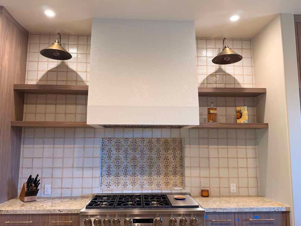 Modern kitchen with white tiled walls, wood shelves, black pendant lights, and a gas stove under a large hood