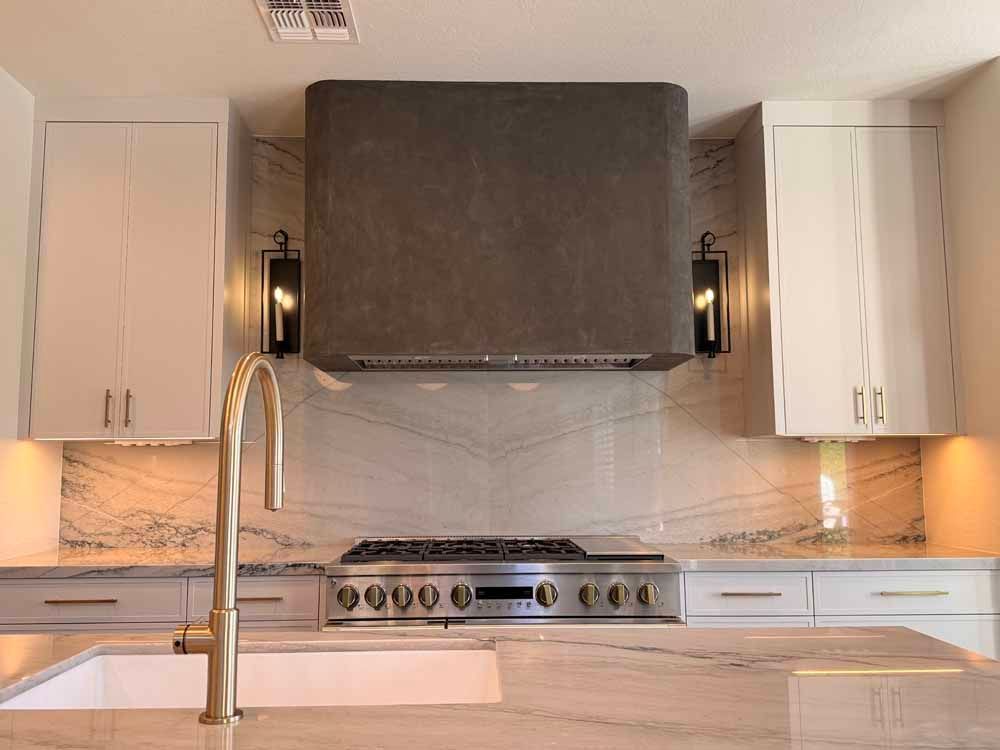 Modern kitchen with white cabinets, stainless range, and large dark vent hood over marble backsplash.