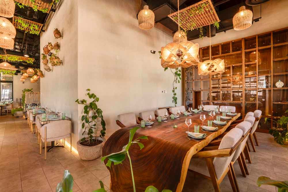 Warm restaurant interior with wooden tables, hanging lights, plants, and shelves of wine glasses