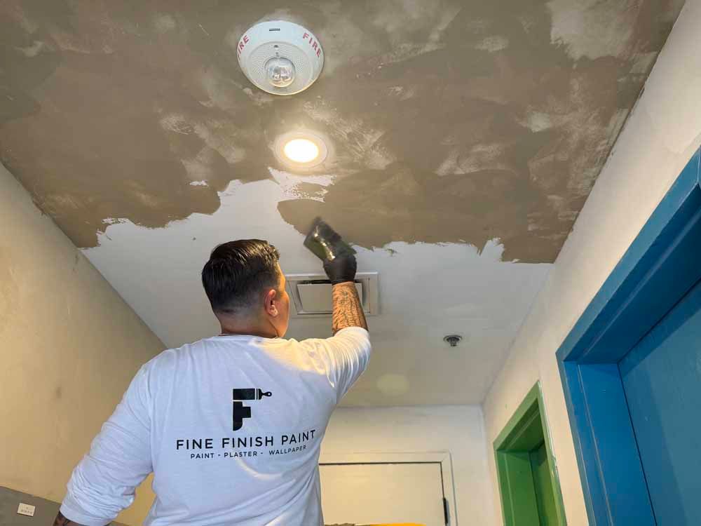 Worker mudding and smoothing a ceiling with a putty knife in a hallway.